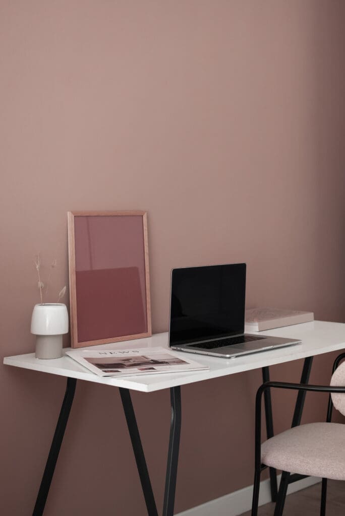 Minimalist blush-toned workspace with a laptop, framed print, and dried florals on a white desk, symbolizing reflection and the quiet process of creativity.